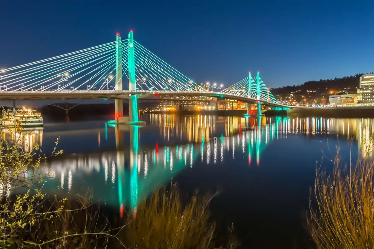 This image highlights a serene suspension bridge over calm water, with reflections and city lights creating a tranquil, picturesque atmosphere. The scene evokes peaceful exploration and a sense of connection to the surrounding locale.
