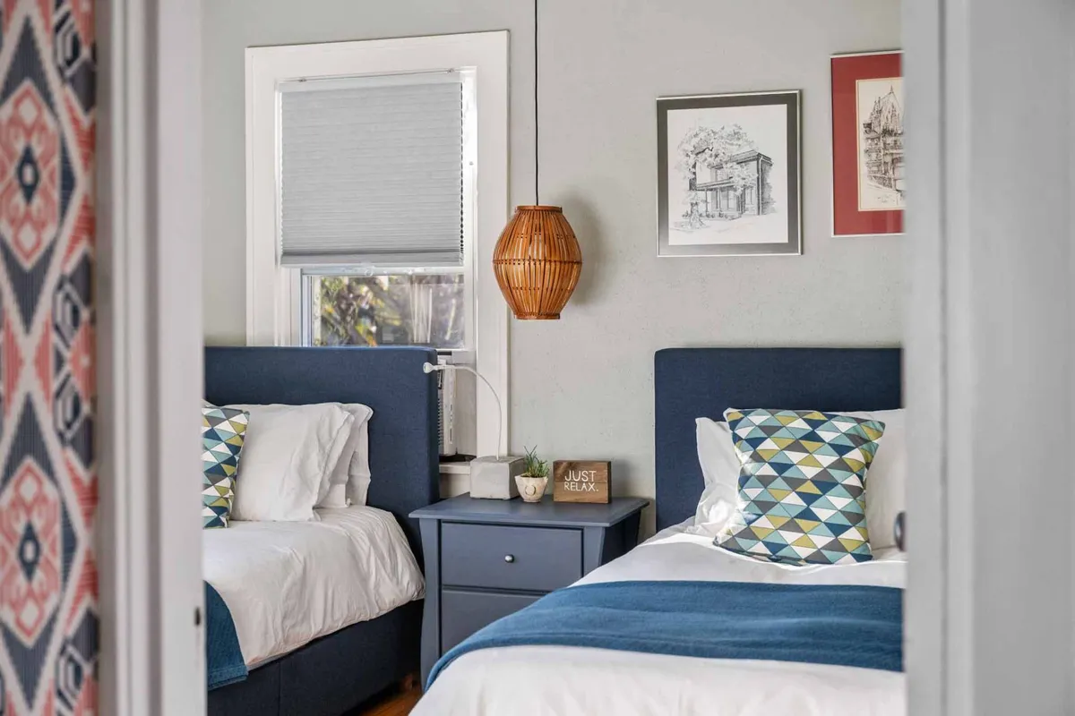 A bright bedroom with two twin beds featuring blue headboards and geometric patterned pillows. Artwork and a pendant lamp add charm, while a decorative sign reading 'JUST RELAX' sets a peaceful tone. The space is designed for both comfort and flexibility.