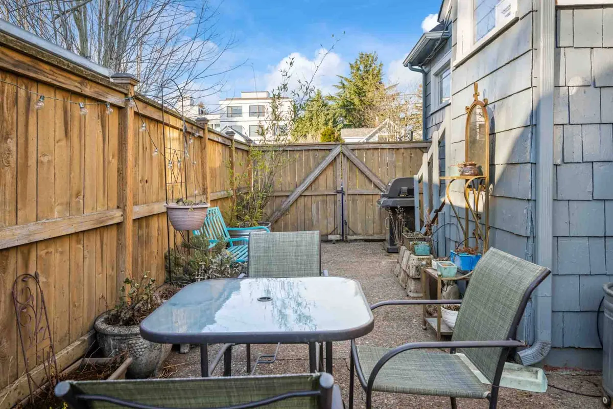 An outdoor dining setup on a patio, featuring a table with blue chairs, a grill, and a fenced garden. The relaxed mood is enhanced by the promise of al fresco dining and celebrations beneath an umbrella or under the open sky.
