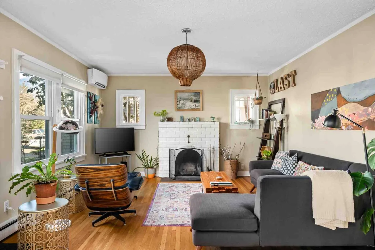 The image showcases a warm and inviting living room anchored by a stylish fireplace, a plush sofa, and a soft rug. Modern artwork and lush green plants add pops of color and personality, creating a space that feels both cozy and vibrant. The overall mood is perfect for relaxing, entertaining, or unwinding with a good book.