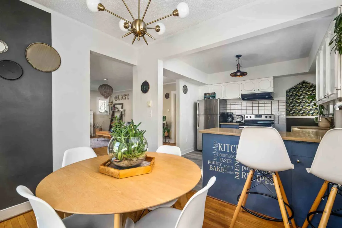 A cozy dining area centered around a round wooden table with a fresh green plant as a centerpiece. The backdrop includes a colorful kitchen backsplash and a modern light fixture overhead, creating a friendly space for meals and conversation.